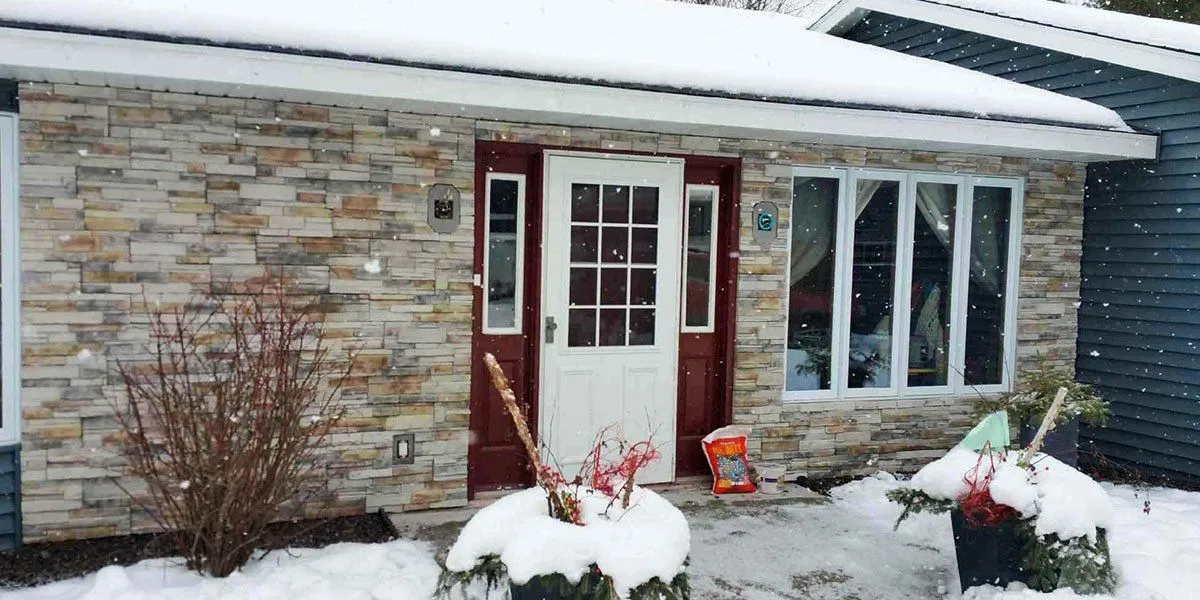 A snowy winter scene in front of a house with stone siding, white door, and potted plants by the entrance.