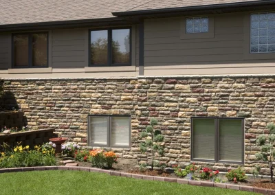 A house with stone siding, flower beds, small bushes, and green grass in the foreground.