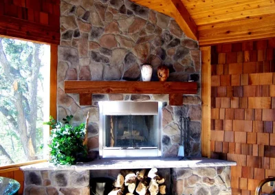 Stone fireplace with wood mantle, logs below, green plant, and vases, in a wood-paneled room.