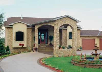 Stone house with arched entry, curved driveway, green lawn, and a fountain in the front yard.