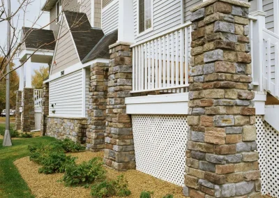 Stone columns and white railings on a porch, with mulch and bushes in the front yard.