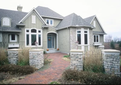 A two-story suburban house with gray siding, stone pillars, and a brick walkway to the front door.