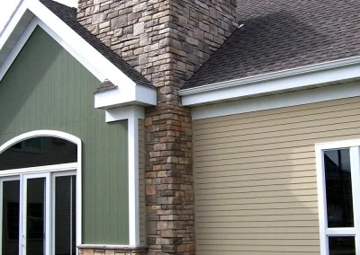House exterior with green and beige siding, stone chimney, large windows, and manicured lawn.