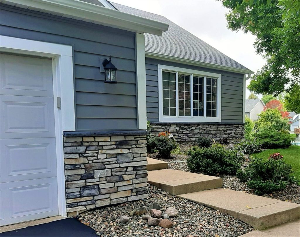 Gray house with stone accents, shrubs, and concrete steps in a landscaped front yard.