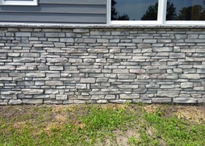 Gray stone veneer wall with grass at the base, under windows and siding on a house exterior.