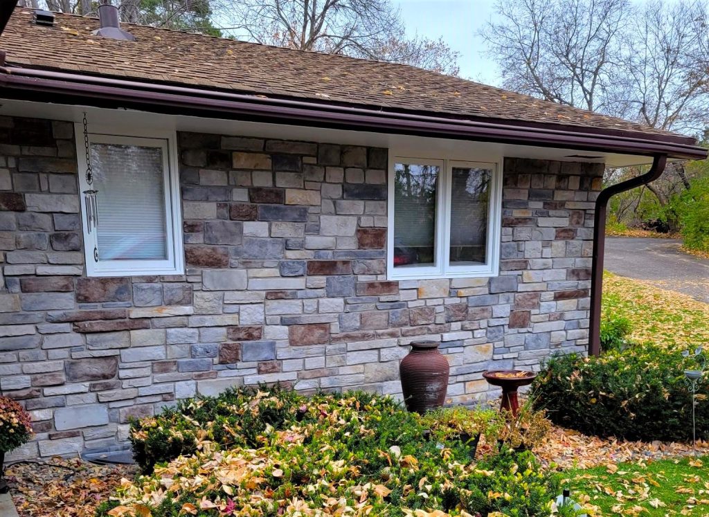 Stone house wall with two windows, an urn, birdbath, and shrubs covered with fallen autumn leaves.