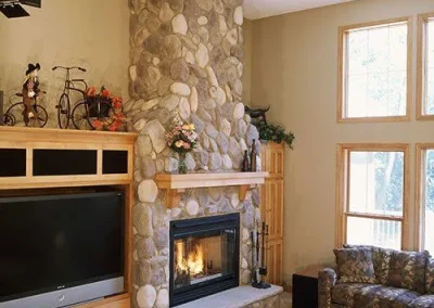 Stone fireplace with fire lit, TV on the left, windows on the right, and a patterned couch in front.