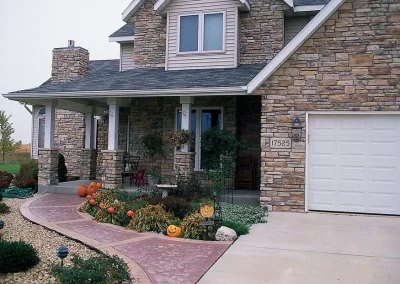 Stone house with front porch, Halloween pumpkins, and fall plants by walkway and garage.