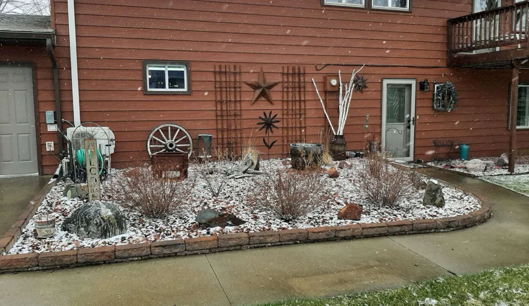 A small, snowy garden with rocks, wagon wheels, and metal stars in front of a red house.
