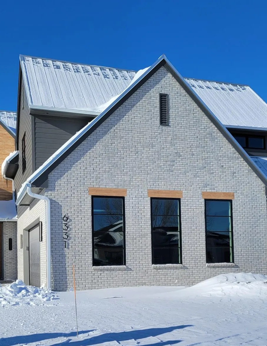 Modern house with gray brick, snow-covered roof, and three large windows in a snowy landscape.