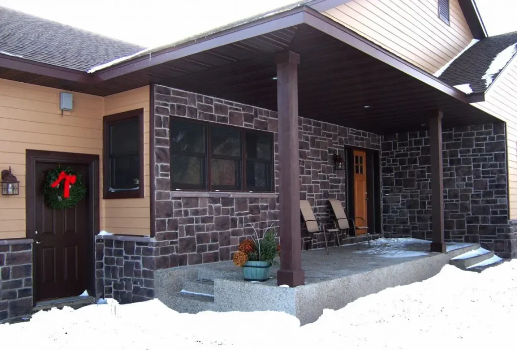 House with stone and siding exterior, two chairs on porch, potted plant, snow on ground, wreath on door.