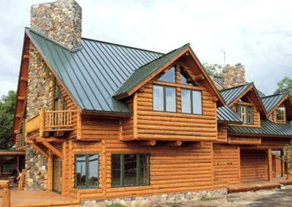 Large log cabin with stone chimneys, green metal roof, big windows, and small balcony on the upper floor.