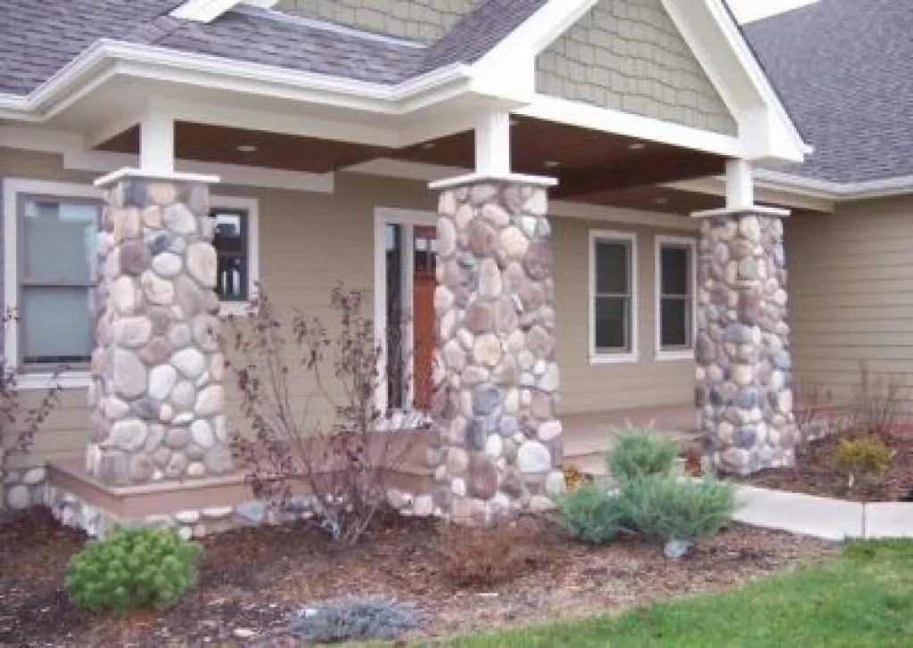 House porch with three large stone pillars, beige siding, white trim, and simple landscaping in front.