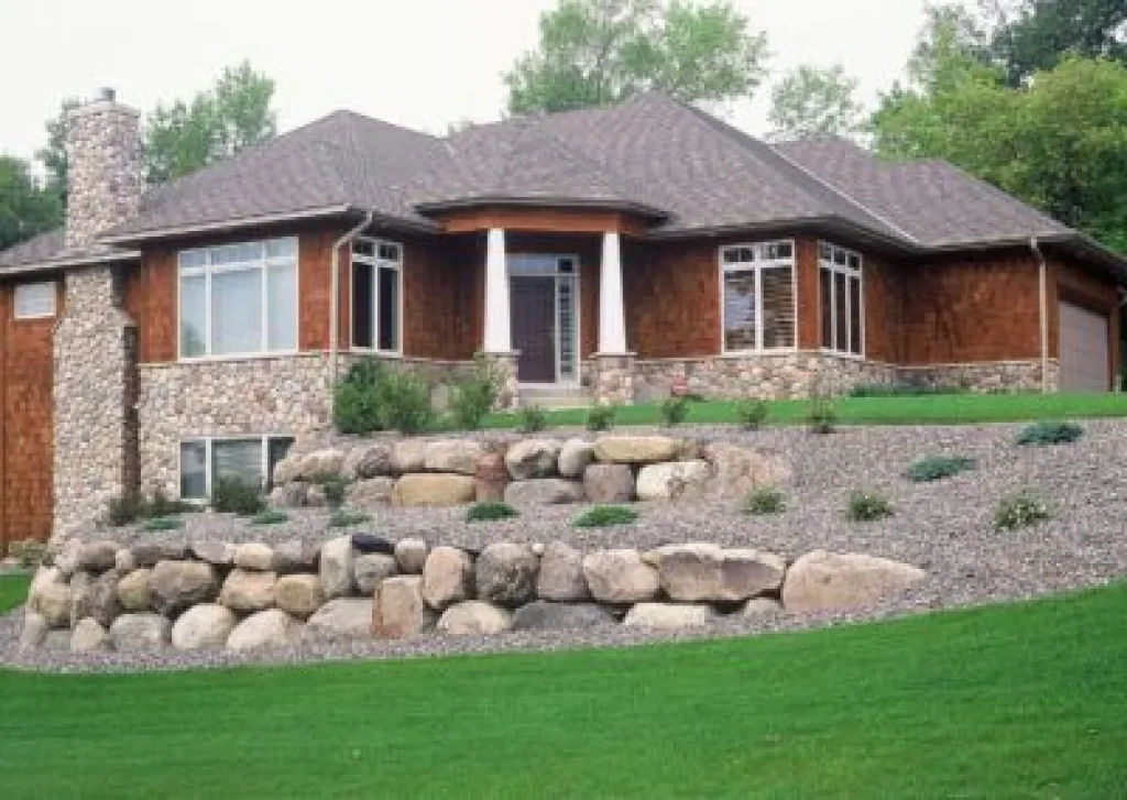 One-story house with stone and wood exterior, large windows, and tiered rock landscaping on a sloped yard.