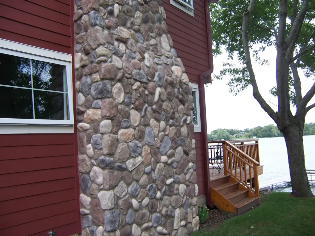 Stone chimney on a red house near a lake, with a wooden deck and trees in the background.