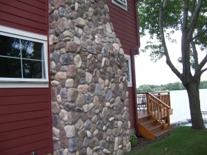 cabin-7 Stone chimney on a red house near a lake, with a wooden deck and trees in the background.