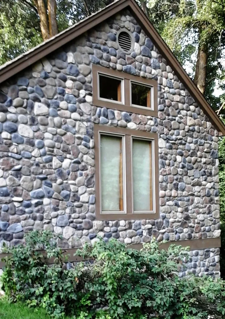 Stone cottage wall with two sets of rectangular windows, surrounded by green bushes and trees.