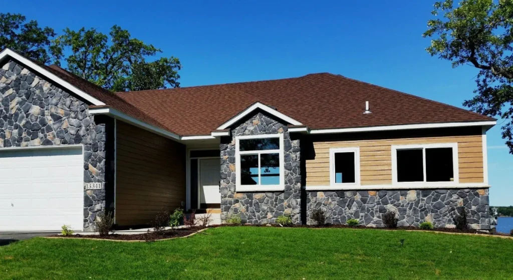 Single-story house with stone and wood siding, brown roof, white garage, and green lawn under blue sky.