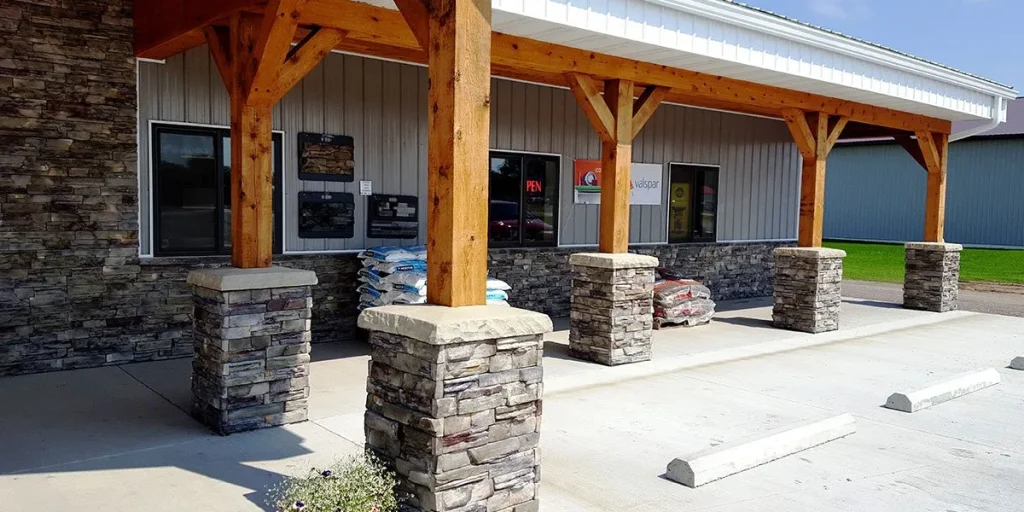 Stone pillars and wooden beams support a porch outside a building with parking spaces in front.