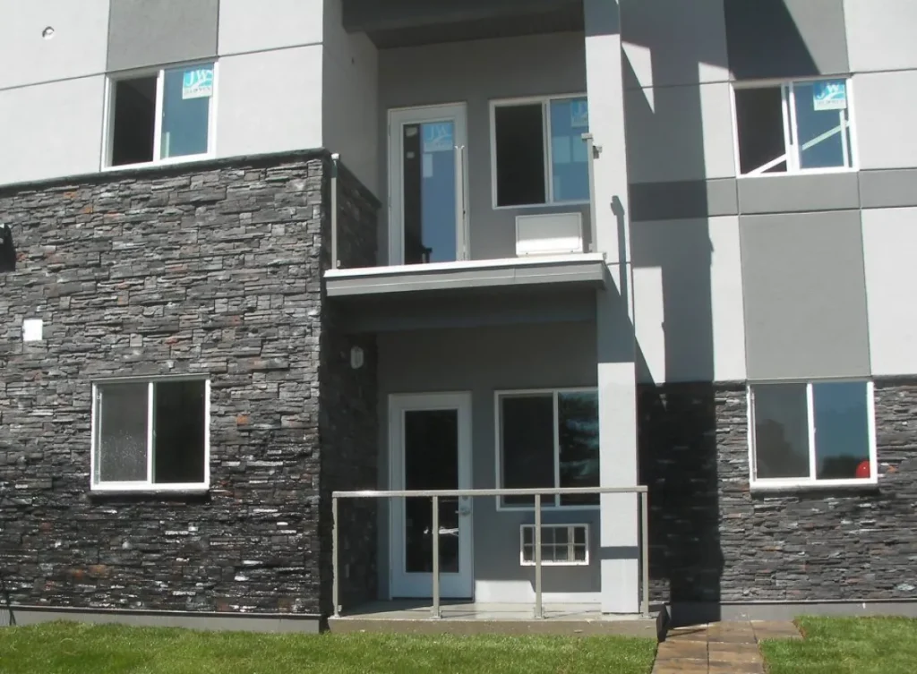 Modern gray apartment building exterior with stone accents, balcony, windows, doors, and grass lawn in front.