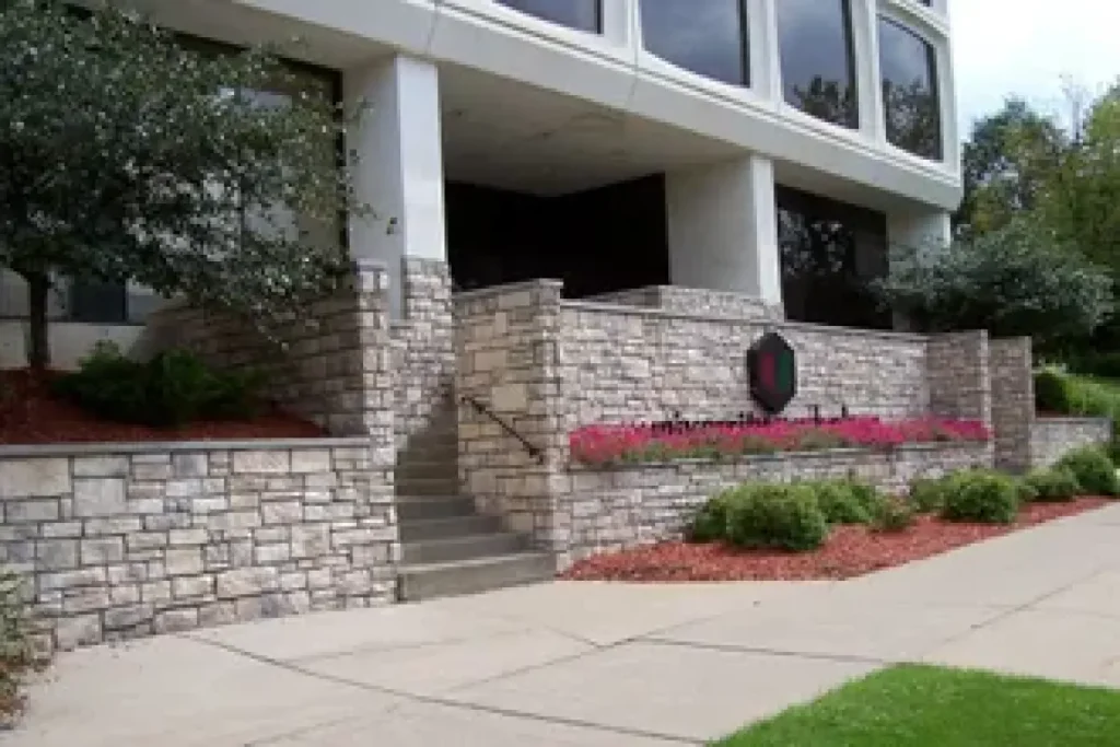Stone building with stairs, bushes, pink flowers, and a sign. Large windows above the entrance.