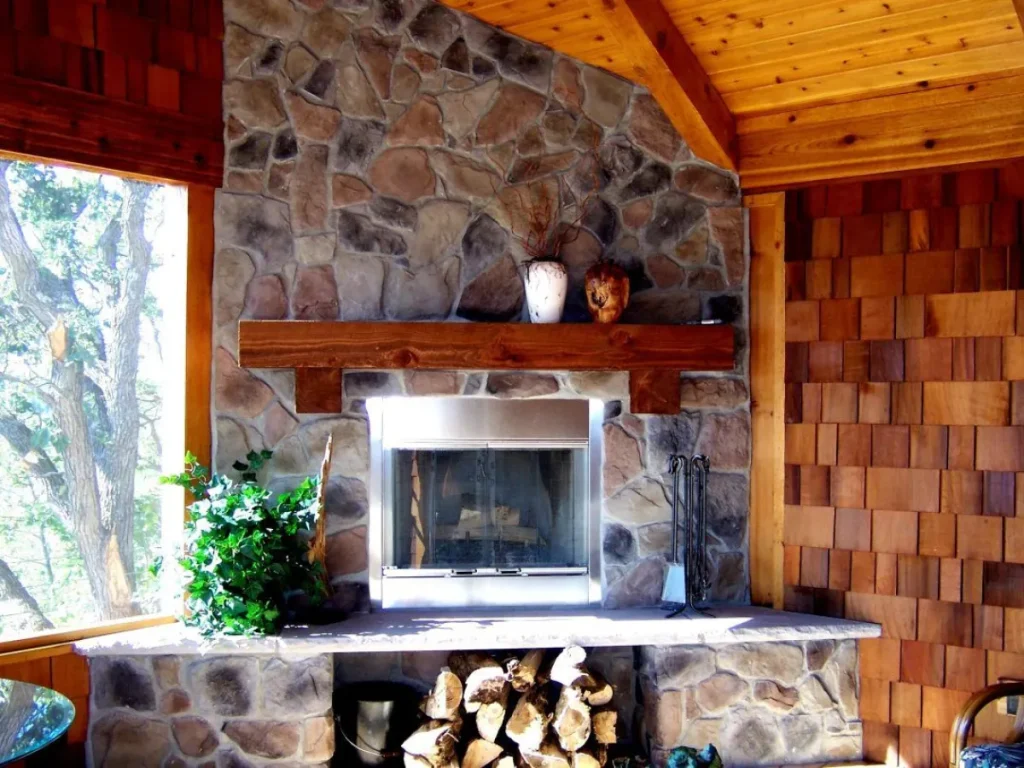 Stone fireplace with a wooden mantel, firewood, potted plant, and wooden ceiling and walls.