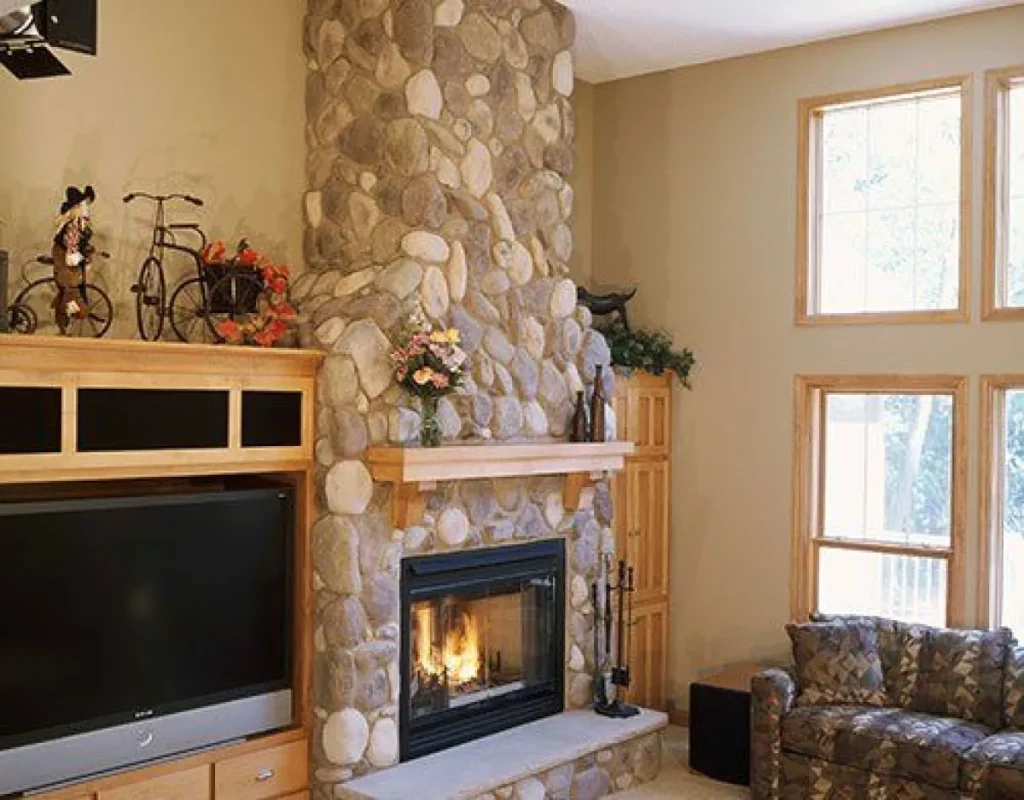 Stone fireplace with a fire, wooden mantel, TV, shelves, and large windows in a cozy living room.