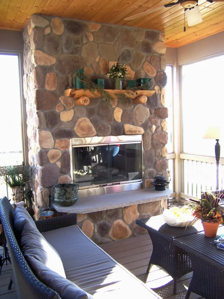 Stone fireplace with a wooden mantel, sofa, and wicker chairs in a sunlit room with wood ceiling.