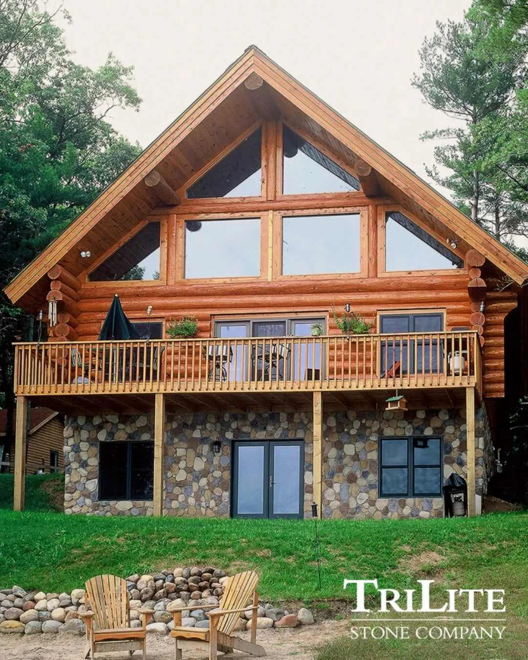 gallery-1 A log and stone cabin with a wooden deck and large windows, surrounded by trees and green grass.