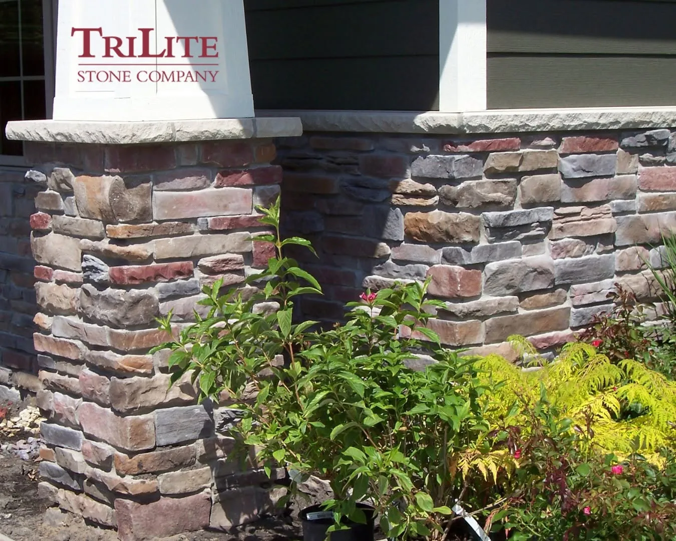 gallery-2 Stone pillars with mixed-tone bricks support a porch, surrounded by green plants and the TriLite Stone Company logo.
