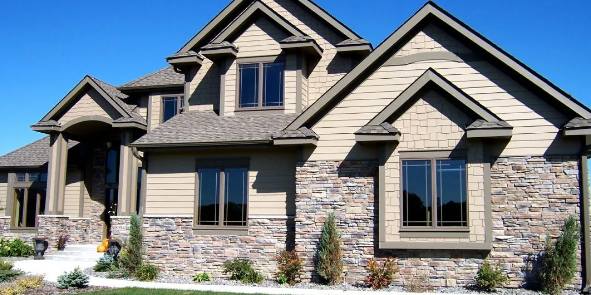 Modern suburban house with tan siding, stone accents, gable roof, and landscaped front yard under clear sky.