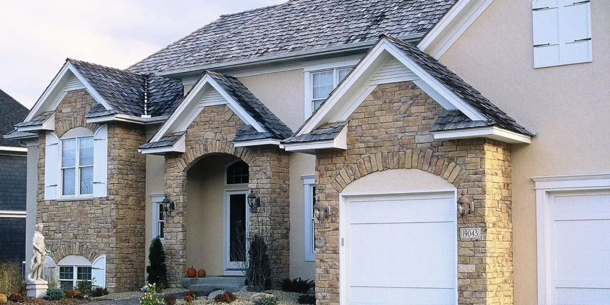 Two-car garage house with stone facade, arched entryway, shingle roof, and small garden in front.