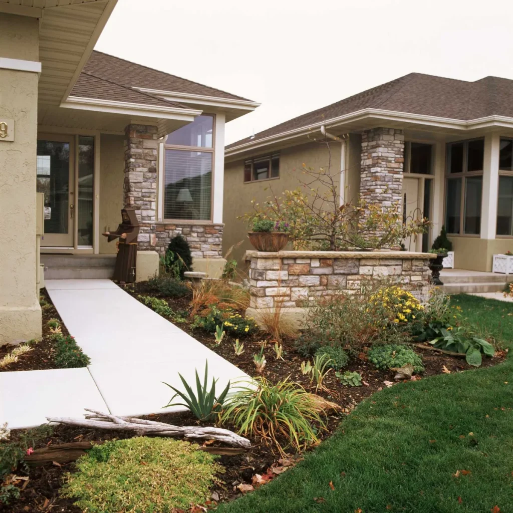 Front yard with garden beds, shrubs, and stone accents next to walkway leading to a house entrance.