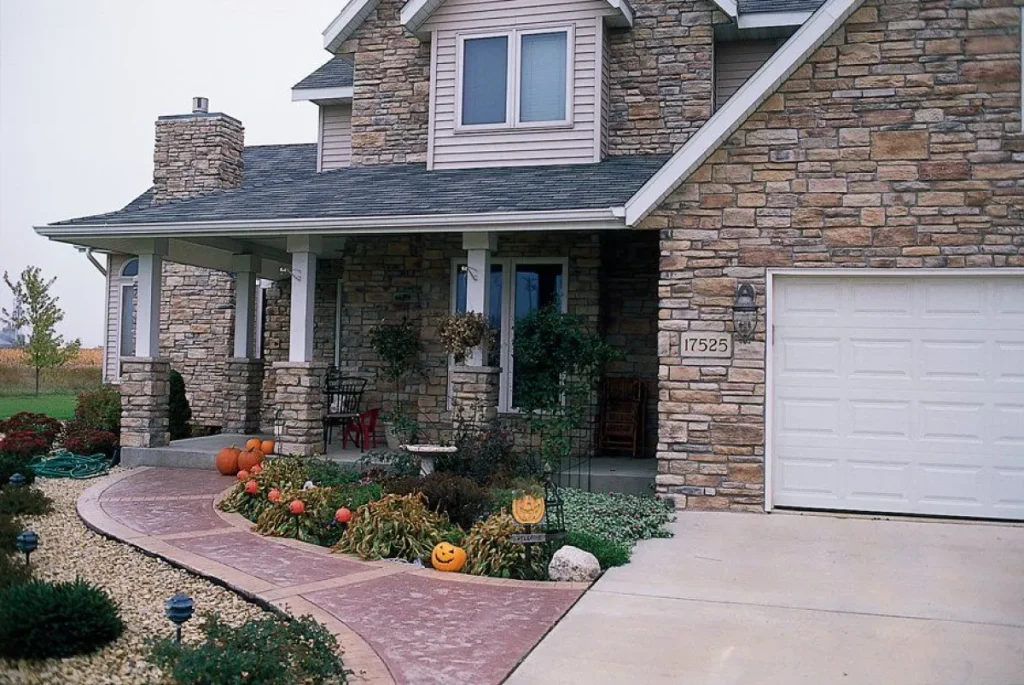 Stone house with porch, carved pumpkins, and fall decorations near walkway. Garage and plants visible.