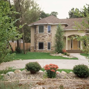 outdoor-4 Two-story house with stone accents, a curved driveway, green lawn, shrubs, and blooming flowers in front.