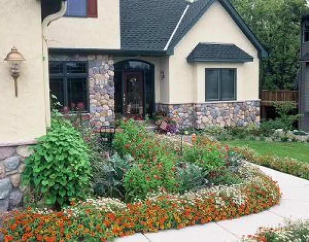 A house with stone accents, a garden with orange and white flowers, and a curved sidewalk in front.