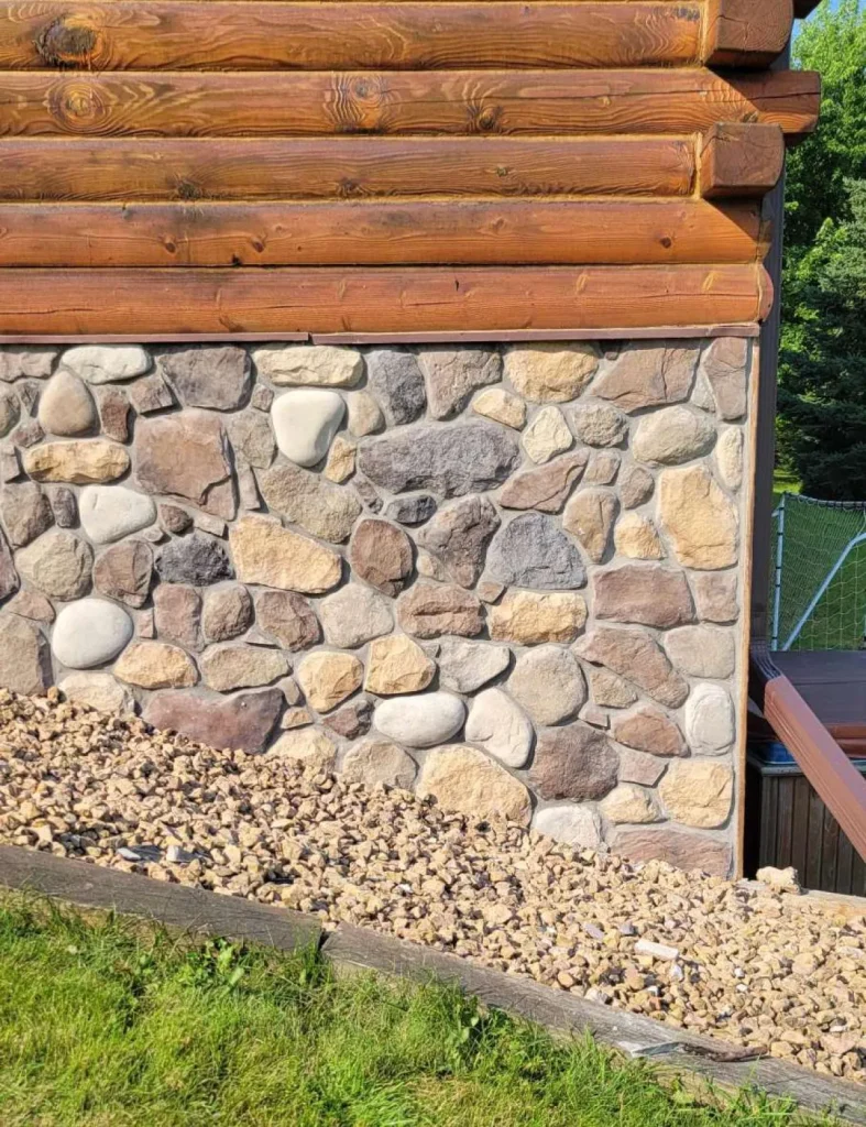 Stone and wood exterior wall with gravel slope and grass in sunlight, bordered by a wooden edge.