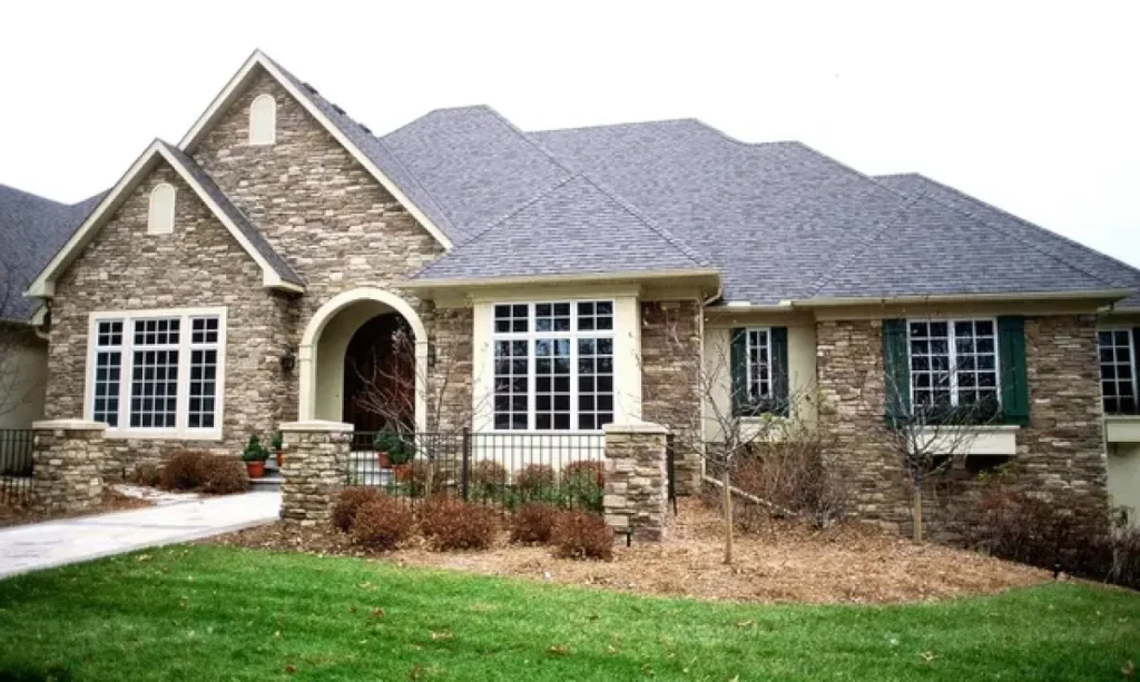 Stone house with arched entry, large windows, gray roof, and landscaped yard with shrubs and green grass.