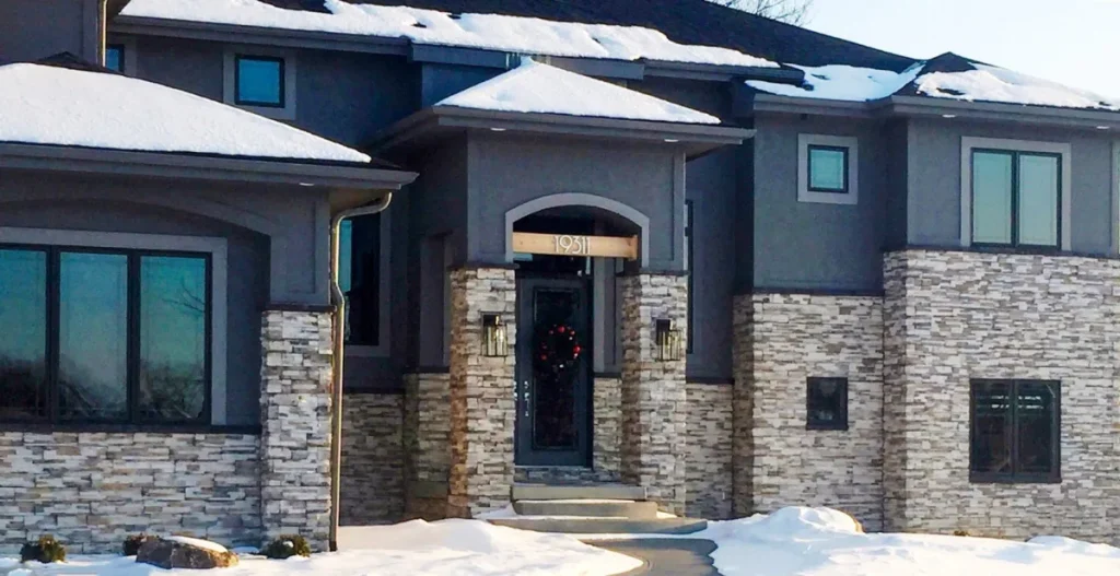 Modern two-story house with stone and dark siding, snow on roof and ground, wreath on front door.