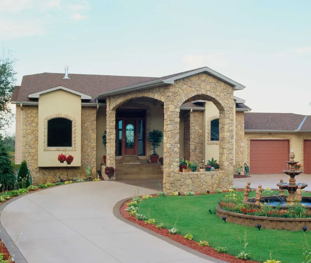 Stone house with arched entry, curved driveway, garden, and a tiered fountain on a lawn.