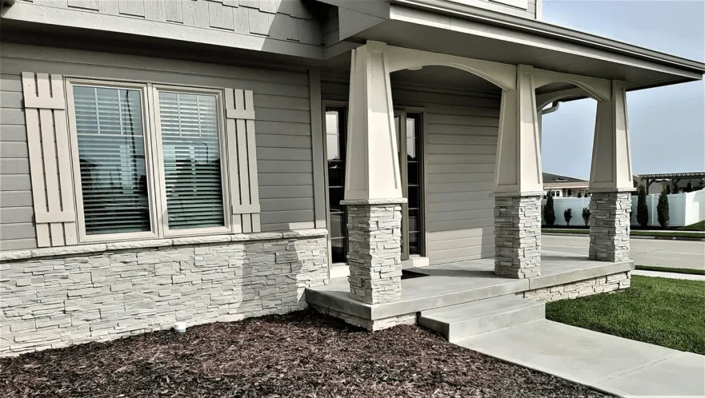 Modern house exterior with stone columns, beige siding, porch steps, and two front windows with shutters.