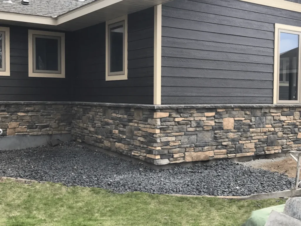 Corner of a house with stone siding, dark brown paneling, and a gravel area next to a patch of grass.