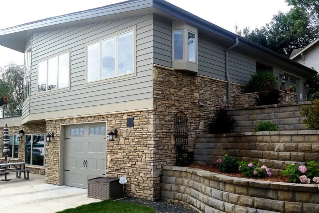 Two-story house with gray siding, stone exterior, garage, and tiered garden beds with flowers.
