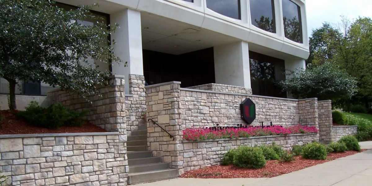 Stone building entrance with stairs, trees, pink flowers, and a black shield logo on the wall.