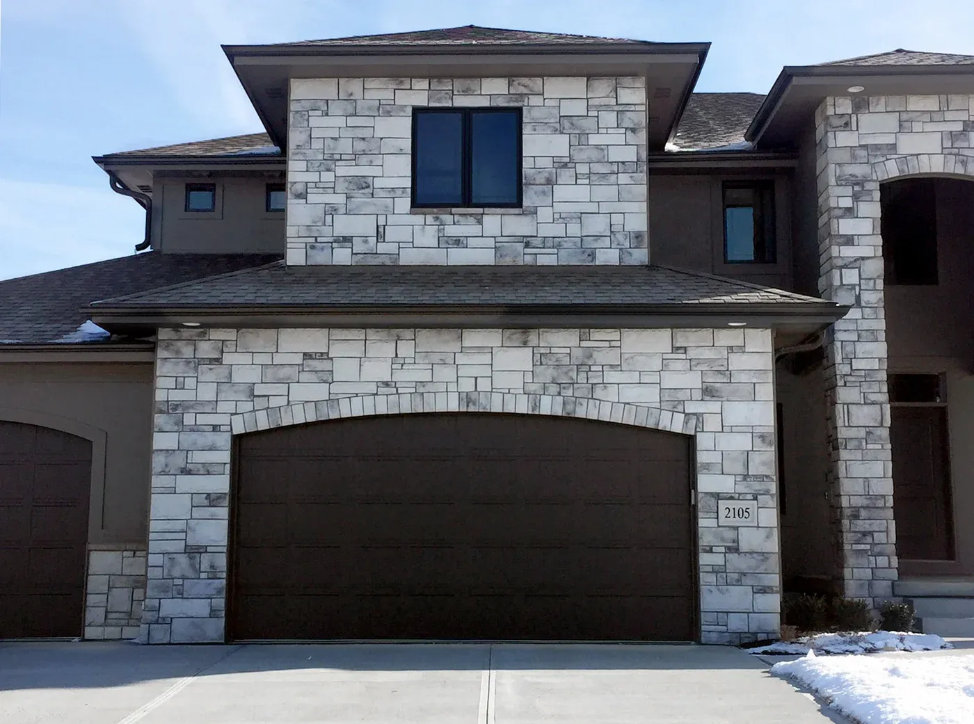 Castlerock Custom Glacier Modern house with light stone facade, brown garage door, and a window above, under a clear blue sky.