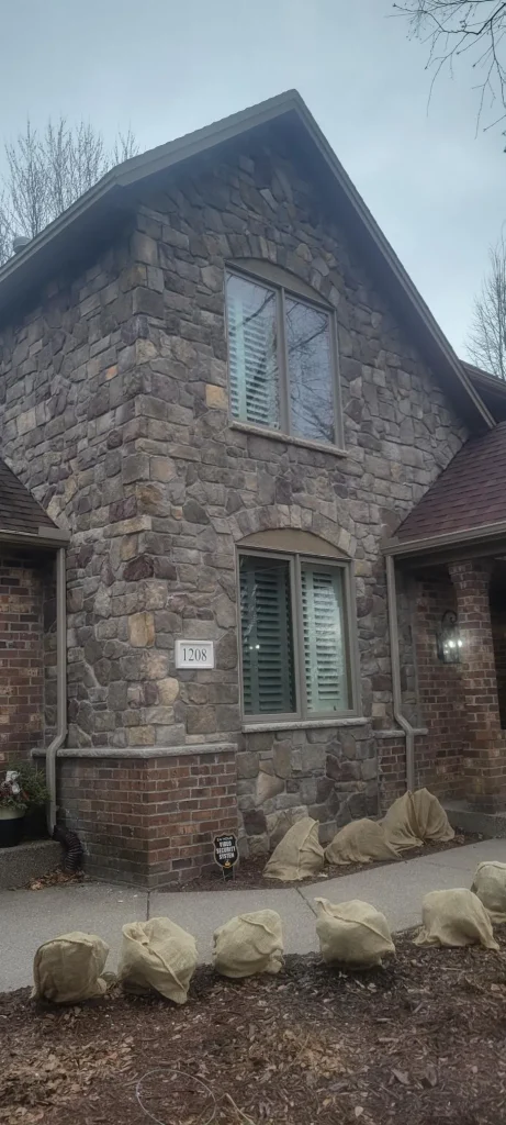 Country-Squire Bucks Country Exterior Stone and brick house with two windows, covered plants by entrance, and address number 1208 visible.