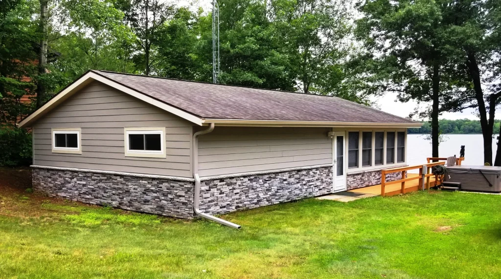 Small gray lakeside cabin with stone siding, surrounded by trees and grass, deck faces the water.