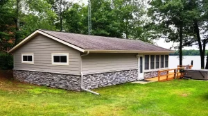 Small gray lakeside cabin with stone siding, surrounded by trees and grass, deck faces the water.