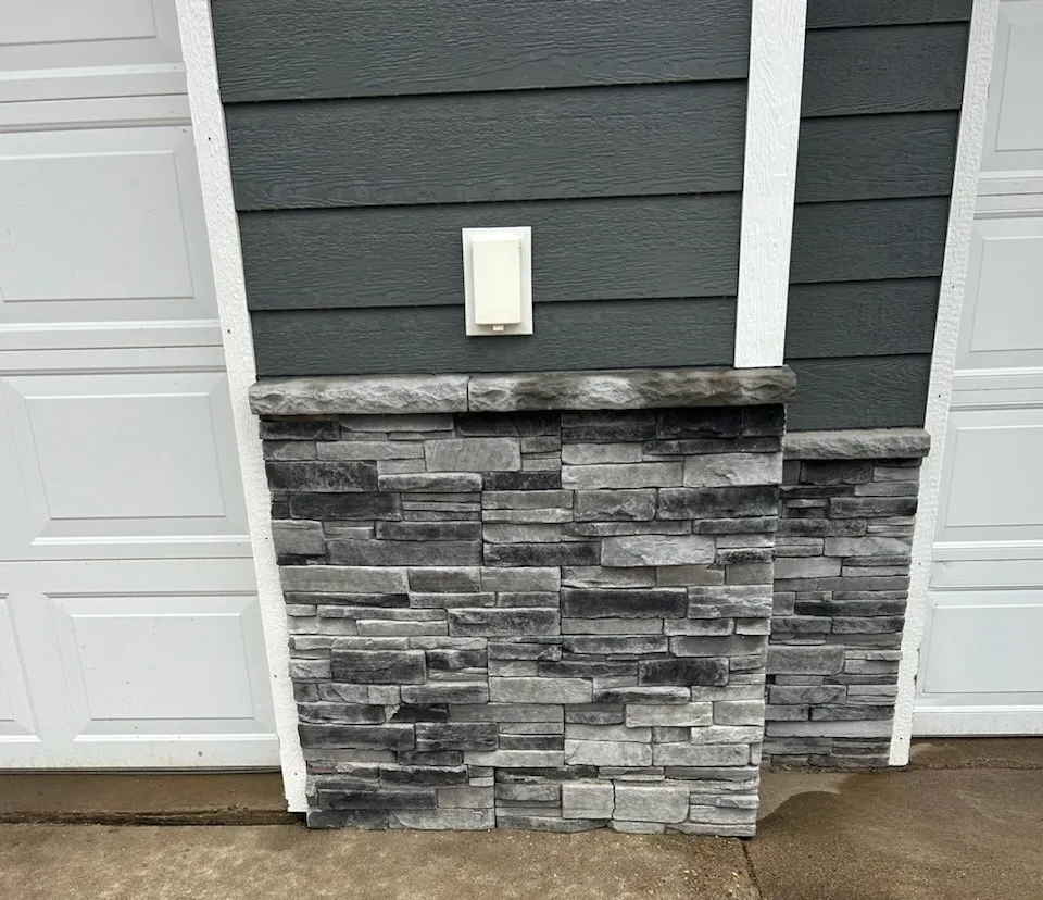 Gray stone veneer on a house exterior, with white trim and a small white outlet cover above the stone.