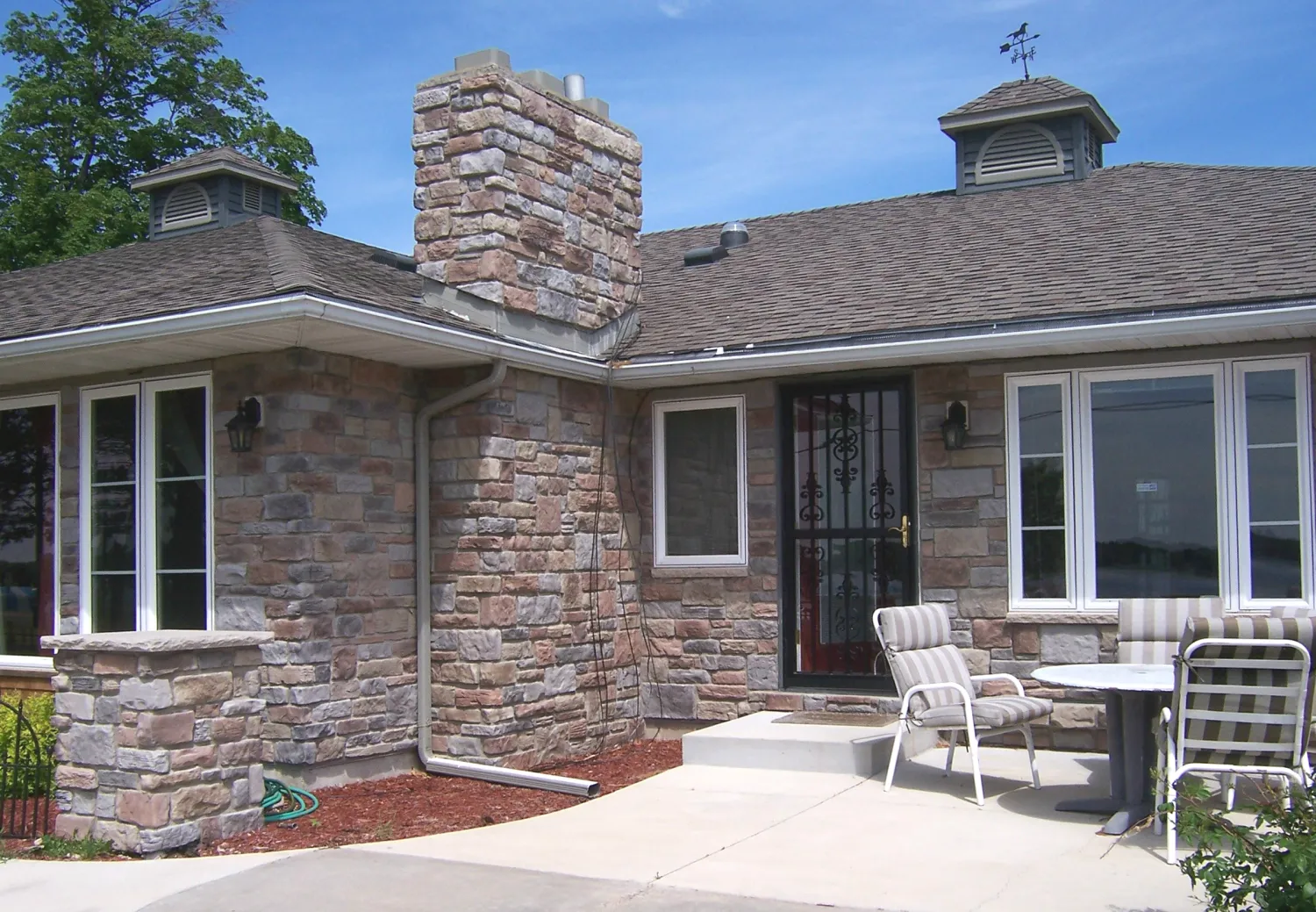 MN Lime Buck_Stein ent 1 Stone house with a chimney, metal door, patio chairs, and table on a sunny day.